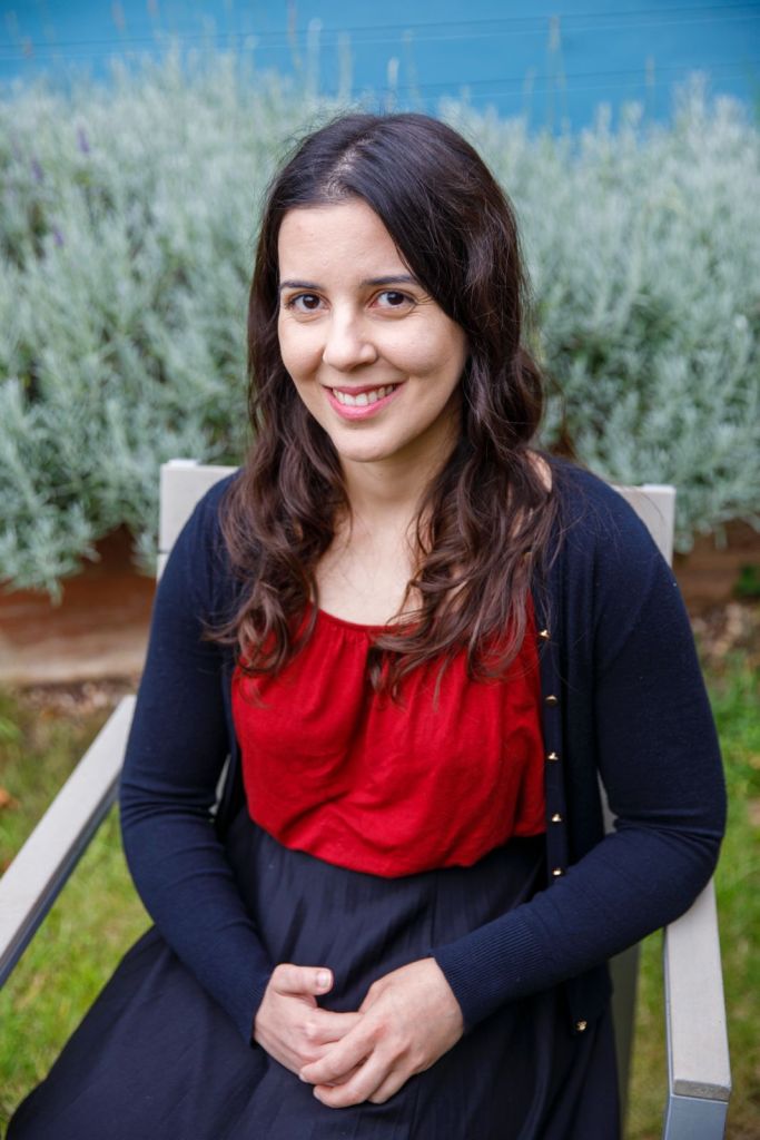 A woman with long, wavy hair and a warm smile is seated outdoors, wearing a red top and a black cardigan. She is sitting in a chair with her hands clasped in her lap, with greenery in the background.